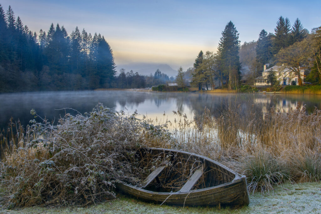 A photograph of Loch Ard, surround by trees, with a white house to the right and a rowing boat in the foreground.