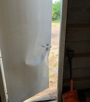 picture shows a door that's slightly ajar with a large dent in it. A sliver of farm field/countryside can be seen through the gap