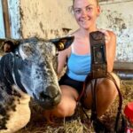 young female farmer holding a black ram harness, crouched in a livestock barn, with a black and white mottled ram on the left, and a red bucket on the right