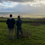 two people standing by a farm gate looking out over farm field with cloudy sky in background