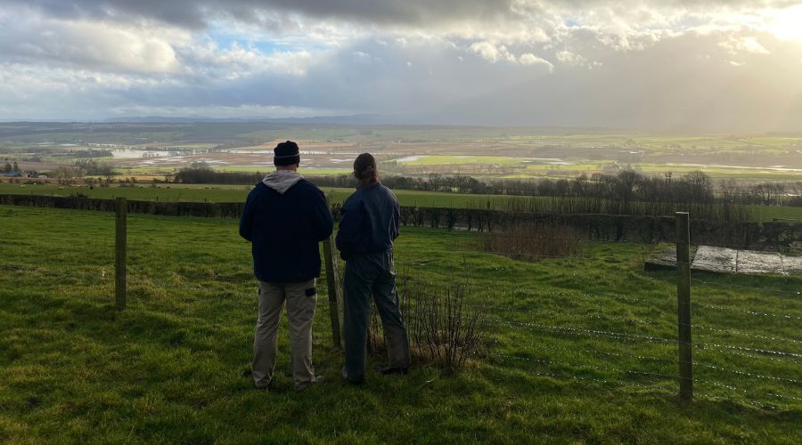 two people standing by a farm gate looking out over farm field with cloudy sky in background