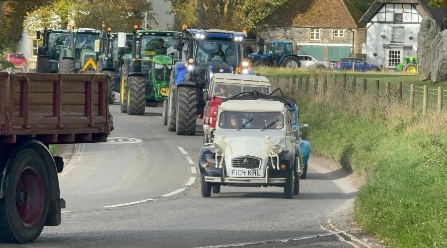 white vintage car with two skeletons attached to the headlights, followed by a line of tractors 