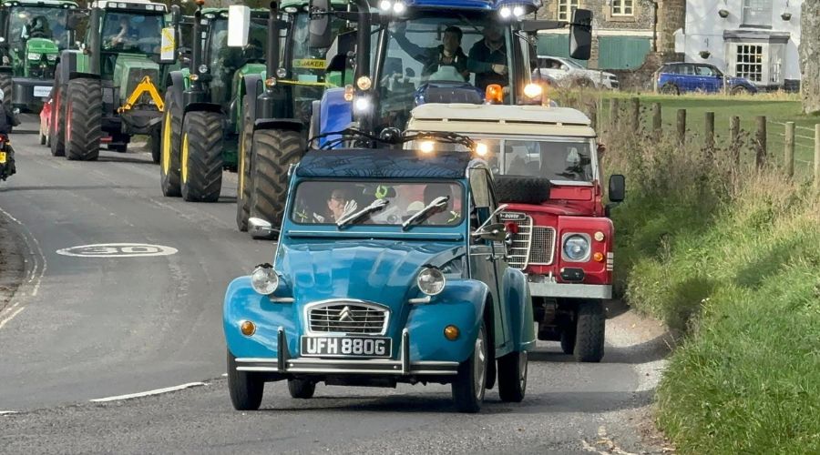 Blue vintage car with skeleton arms for windshield wipers, followed by a line of tractors driving in tractor run