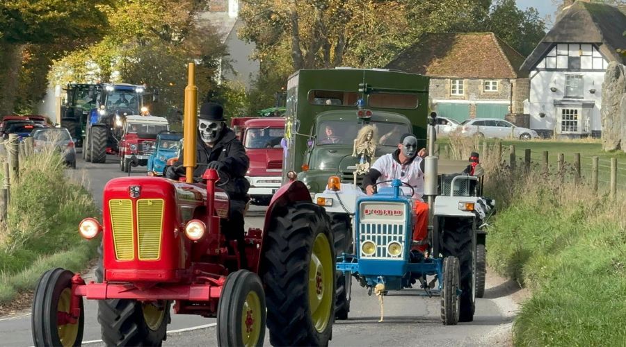 Red vintage tractor with a driver in black wearing a top hat and mask, followed by a blue ford vintage tractor with a masked driver, and a line of other decorated cars and tractors following behind