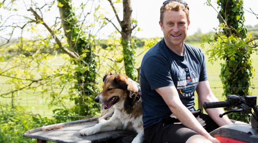 Dan Pritchard, smiling, sits on an ATV with a collie dog behind him and trees and field in the background. 