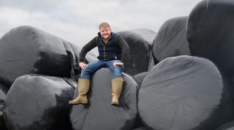 Kaleb Cooper sat smiling on top of a pile of bales in black plastic wrap.