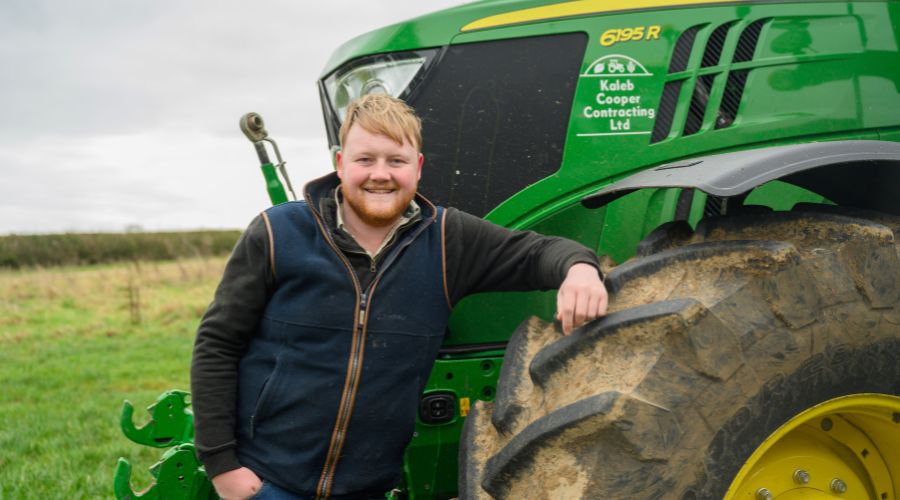 Kaleb Cooper standing in front of a John Deere tractor with Kaleb Cooper Contracting Ltd written on it