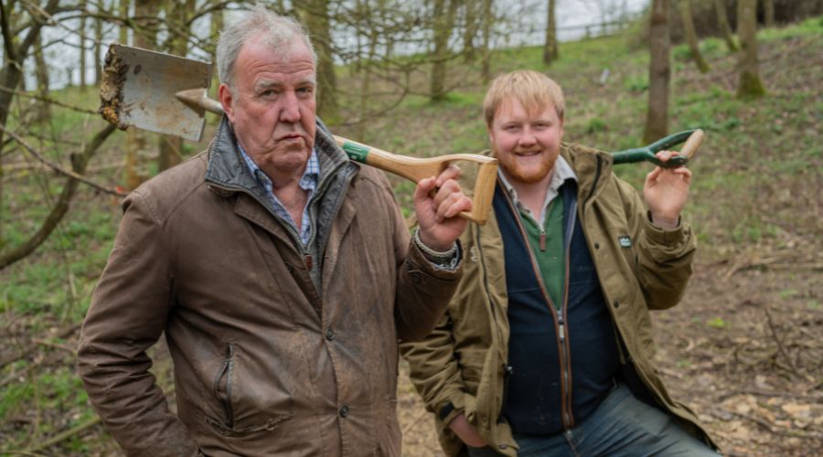 Jeremy Clarkson and Kaleb Cooper standing in the woods, each carrying a shovel over their shoulders.