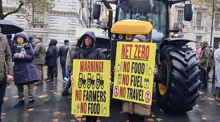 crowd of farmers with large tractor in the centre. Two farmers in front of the tractor are holding yellow banners saying 'warning, no farmers no food' and 'net zero, no food, no fuel, no travel'