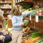 father and daughter in a farm shop - father crouches on the floor while young daughter reaches up to a shelf full of produce including carrots, onions and potatoes