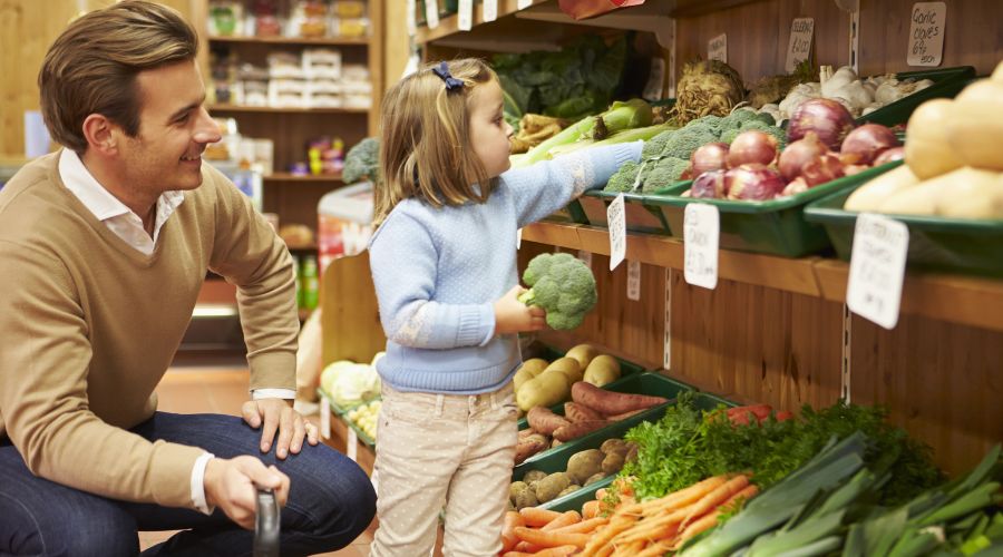 father and daughter in a farm shop - father crouches on the floor while young daughter reaches up to a shelf full of produce including carrots, onions and potatoes