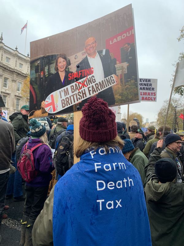 picture of crowd of farmers holding banner showing Rachel Reeves with text saying 'I'm shafting British farming'. A woman in the foreground is shown from the back with a blue flag draped around her saying 'family farm death tax' 