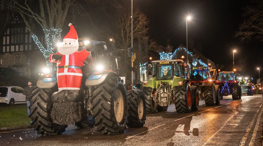 Line of festively decorated tractors driving down a road with an inflatable Santa on the front of the first tractor 