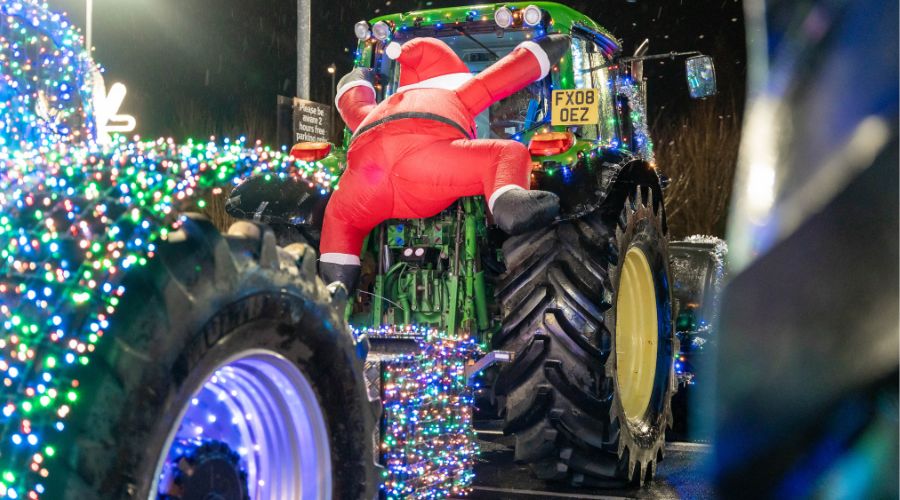 back of John Deere tractor with an inflatable Santa hanging onto the back. Another tractors wheels, covered in lights, are visible behind 