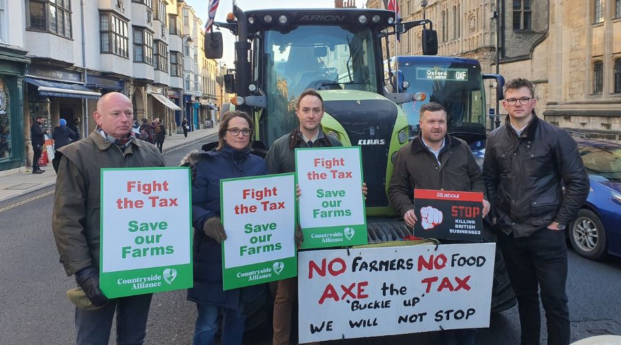 group of people standing in front of a tractor at Oxford farmers protest, holding placards saying fight the tax save our farms 