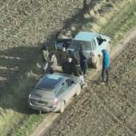 group of hooded masked people standing in an arable field with two cars