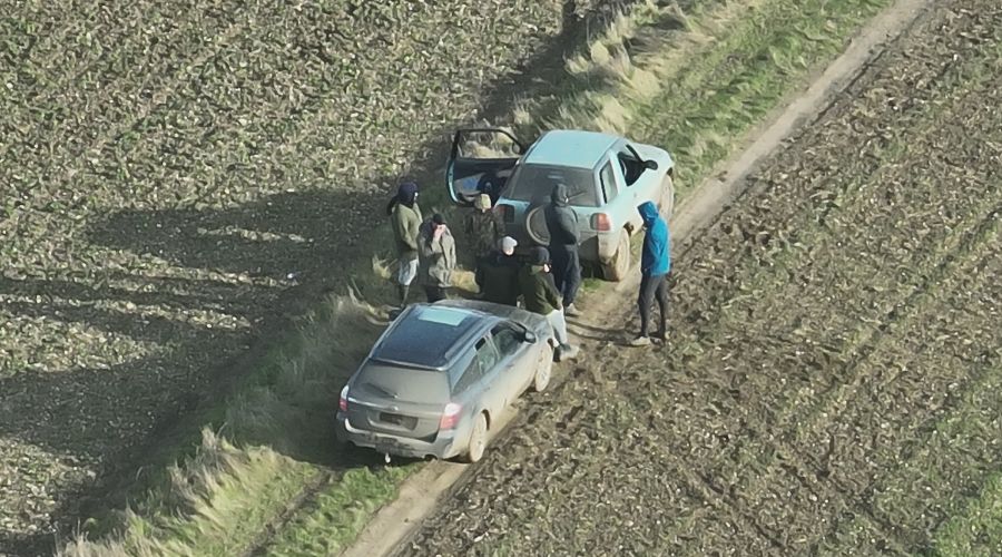 group of hooded masked people standing in an arable field with two cars