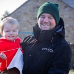 tom Carlisle holding a young child standing in front of a farm building