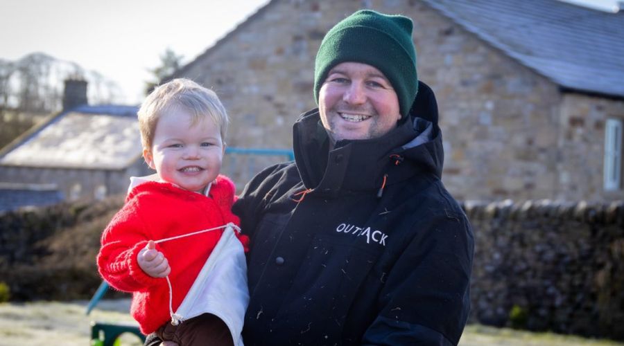 tom Carlisle holding a young child standing in front of a farm building