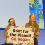 two protesters on stage at the NFU conference wearing carrot masks and holding a sign saying Root for the planet go vegan